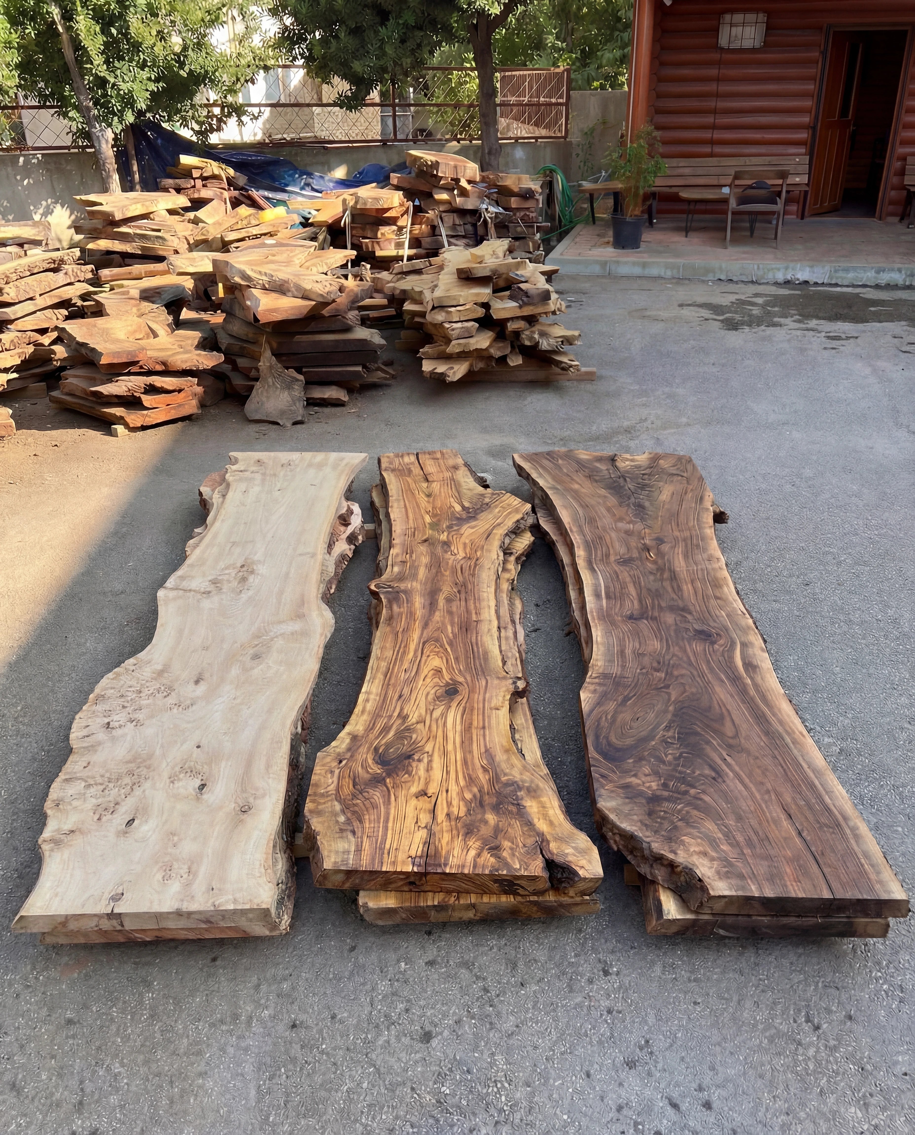 Three wooden planks with different finishes on a concrete surface, with stacks of wood in the background.